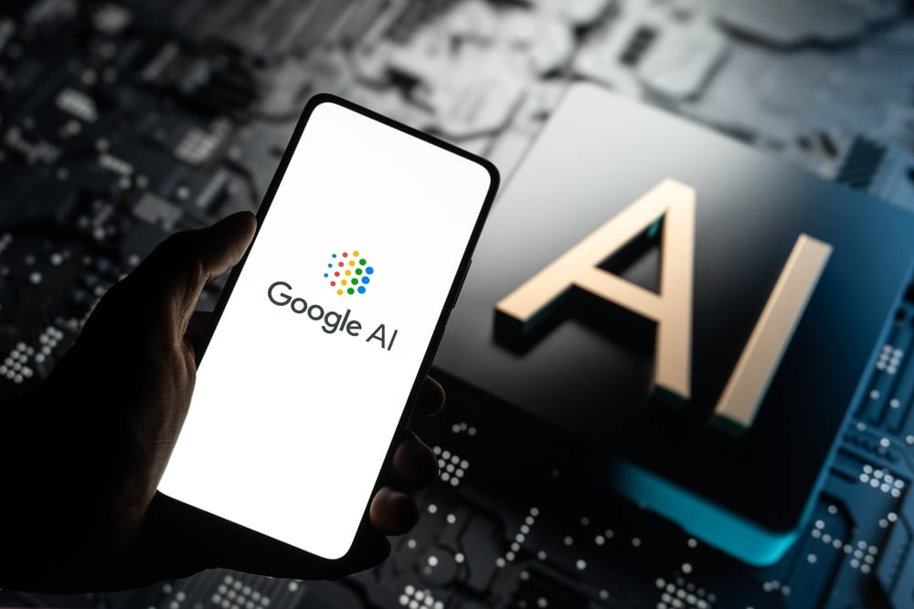 Hand holding a smartphone displaying the Google AI logo with a large AI symbol in the blurred background, suggesting a tech environment.