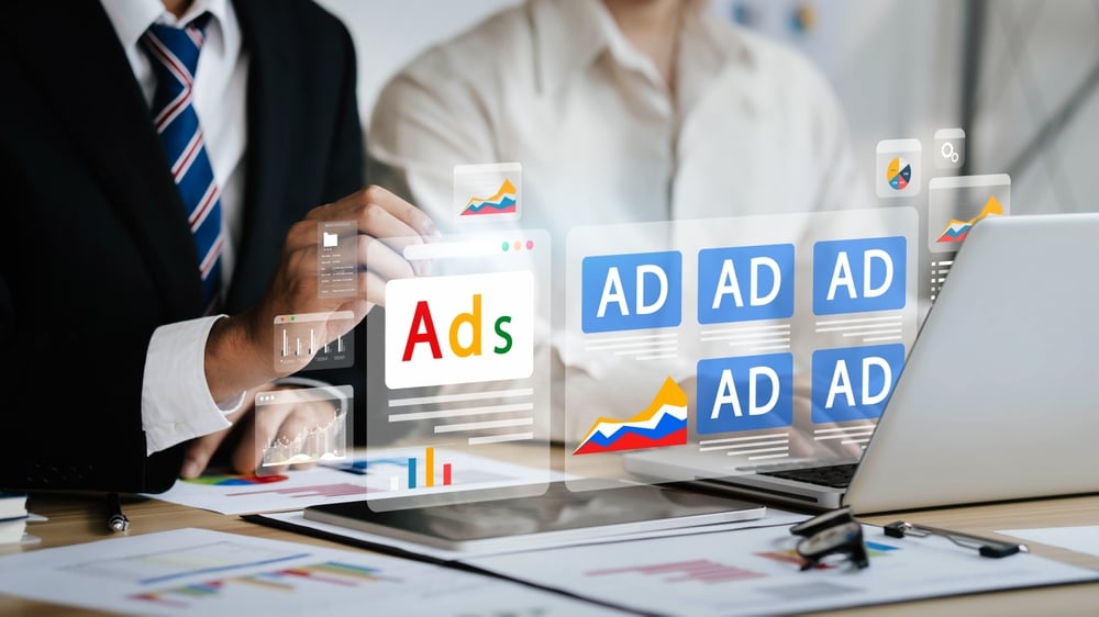 Two professionals reviewing digital ads on a laptop with floating ad icons and graphs from a meeting.