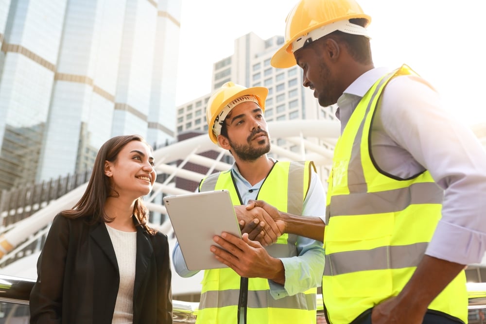 Two construction workers in yellow hard hats and safety vests shake hands on a city construction site, while a smiling woman holds a tablet nearby.