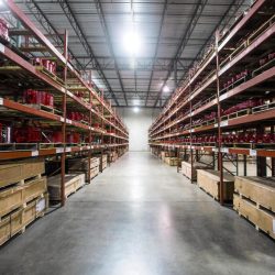 Long Warehouse Aisle with Tall Red Metal Racks Wooden Pallets and Red Items Stored on Both Sides Lit by Overhead Lights at the Ceiling