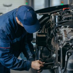 Auto Technician in Blue Coveralls Leaning over an Open Car Hood Using a Tool on the Engine Corridorshaped Components Nearby