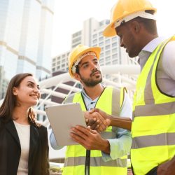 Two construction workers in yellow hard hats and safety vests shake hands on a city construction site, while a smiling woman holds a tablet nearby.