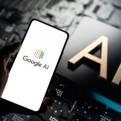 Hand Holding a Smartphone Displaying the Google Ai Logo with a Large Ai Symbol in the Blurred Background Suggesting a Tech Environment