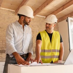 Male Contractor and Engineer Discussing Blueprint Using Laptop During Building Construction